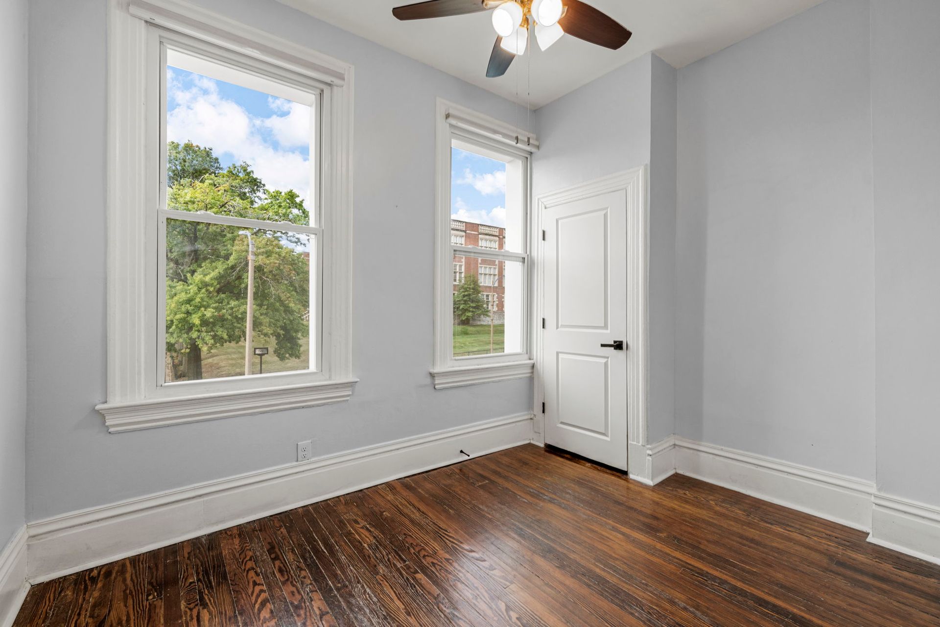 Empty bedroom with hardwood floors, two windows with a view, and a white door.