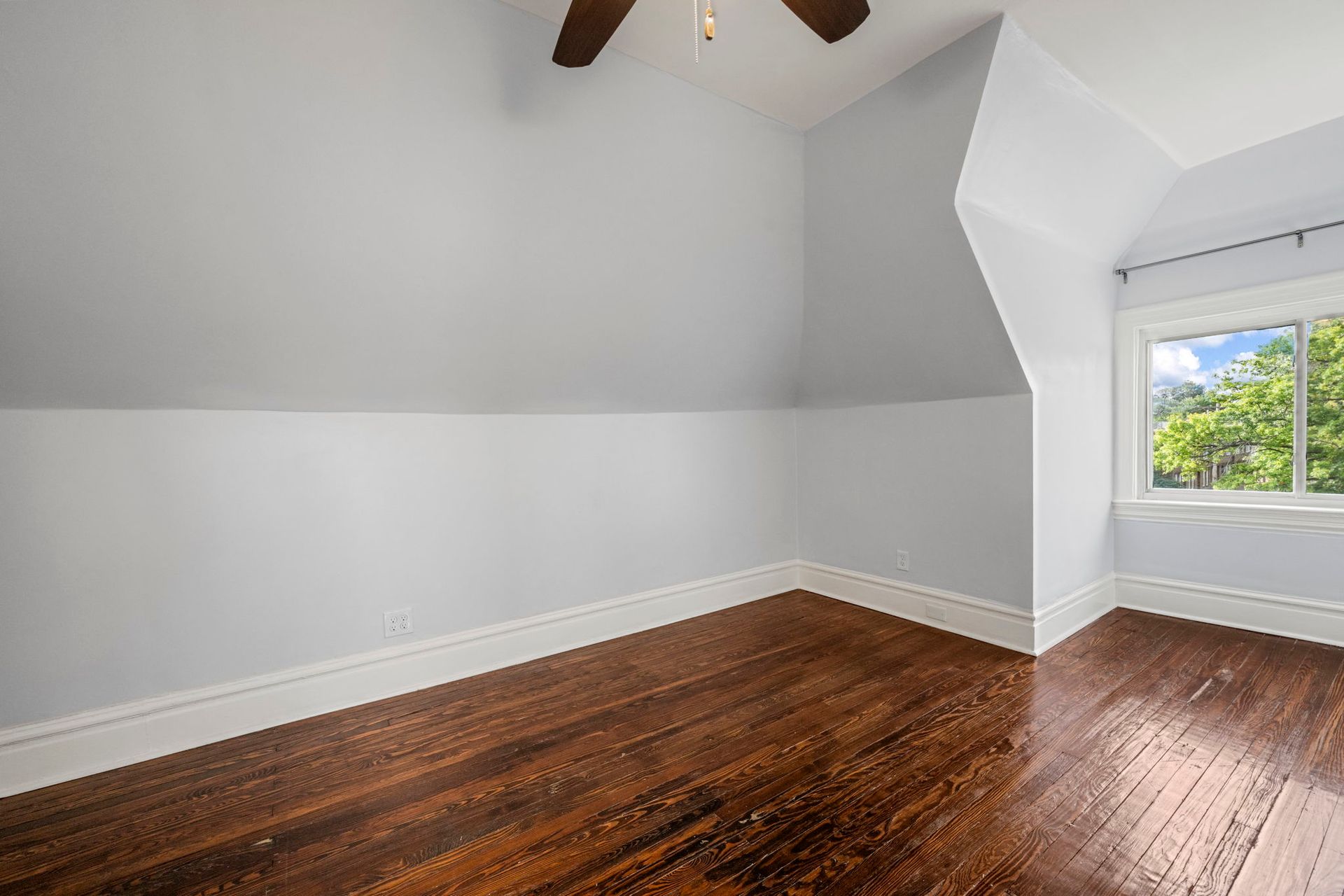 Empty room with light blue walls, dark wood floors, and a window with a view of trees.
