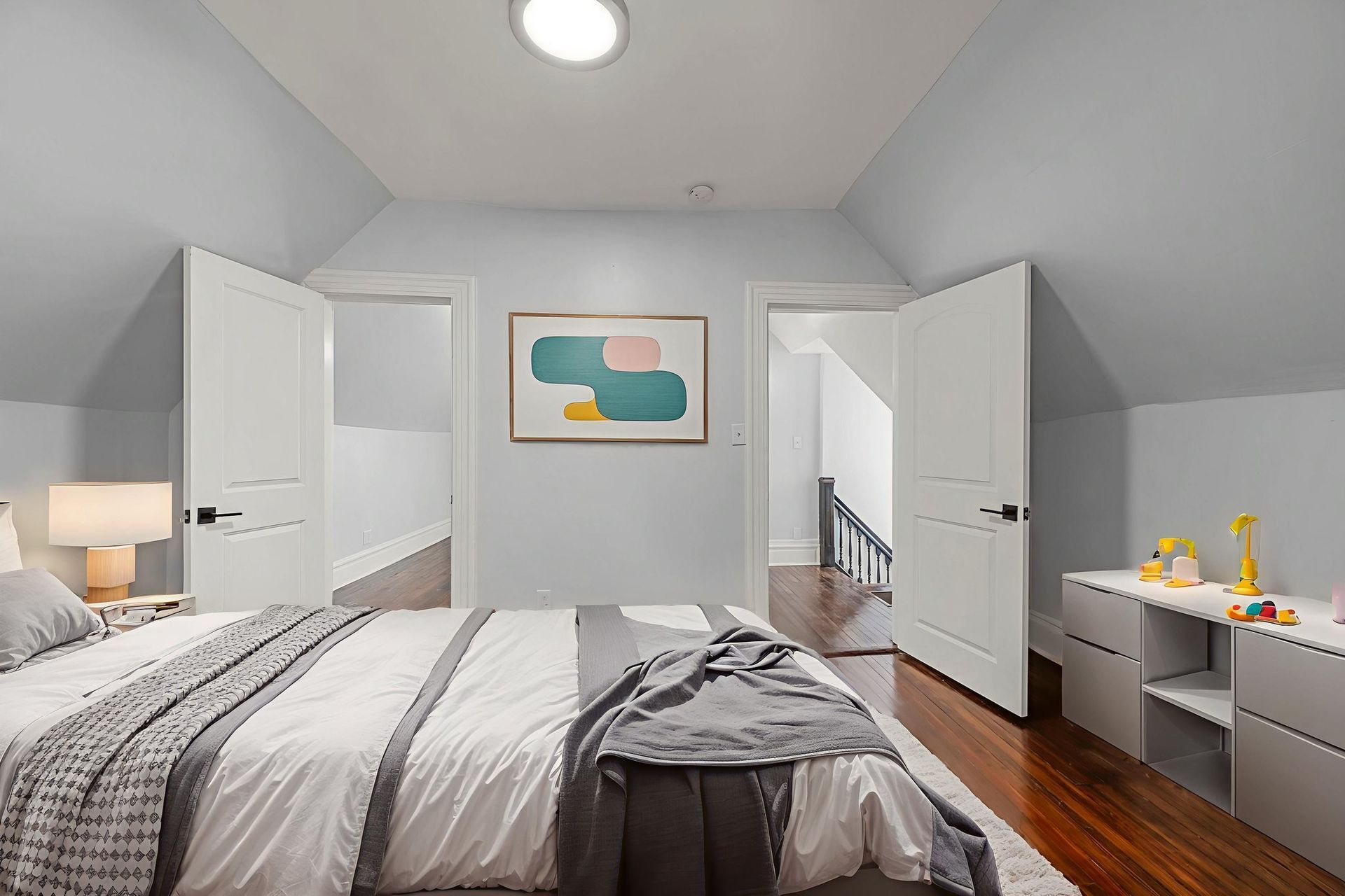 Bedroom with bed, doors, artwork, gray and white bedding, and wooden floors.