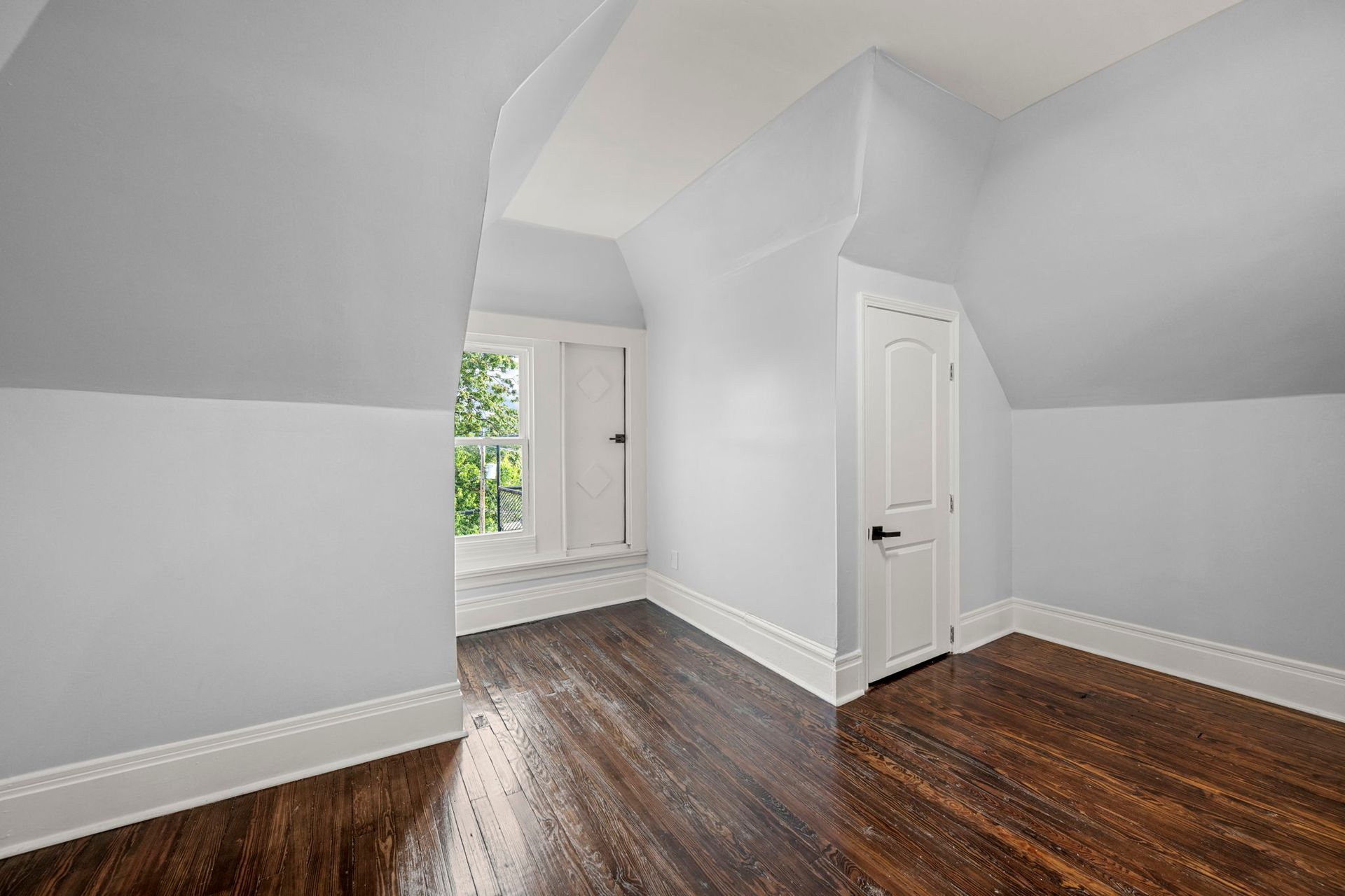 Empty attic room with gray walls, white trim, dark wood floor, window, and door.