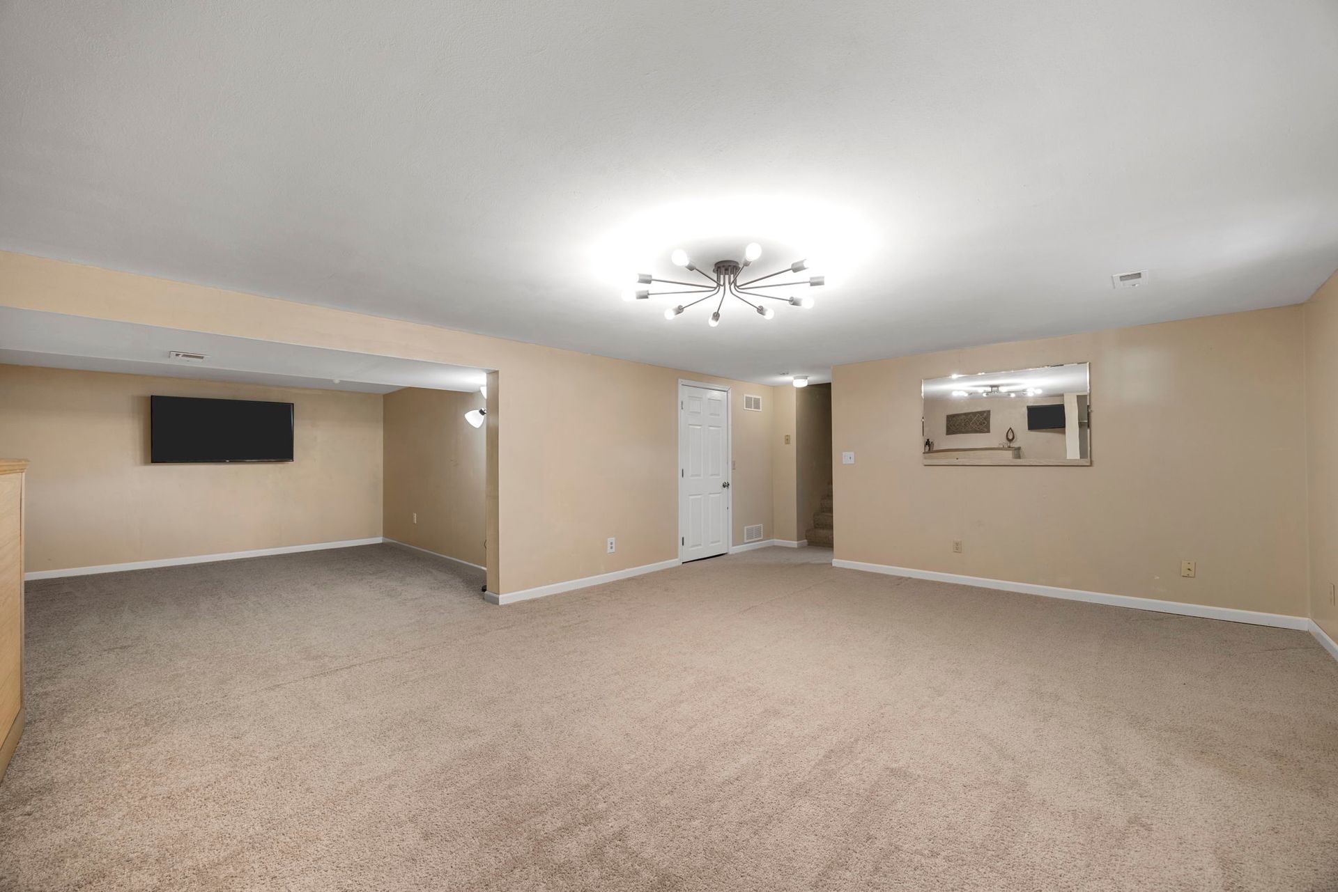 Empty beige-walled room with carpet, ceiling light, TV on wall, doorway, and a window with mirror.