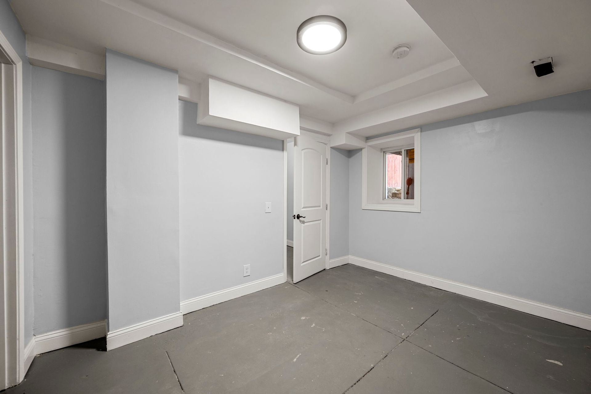 Empty room with light blue walls, white trim, and gray flooring. A closed white door is centered.