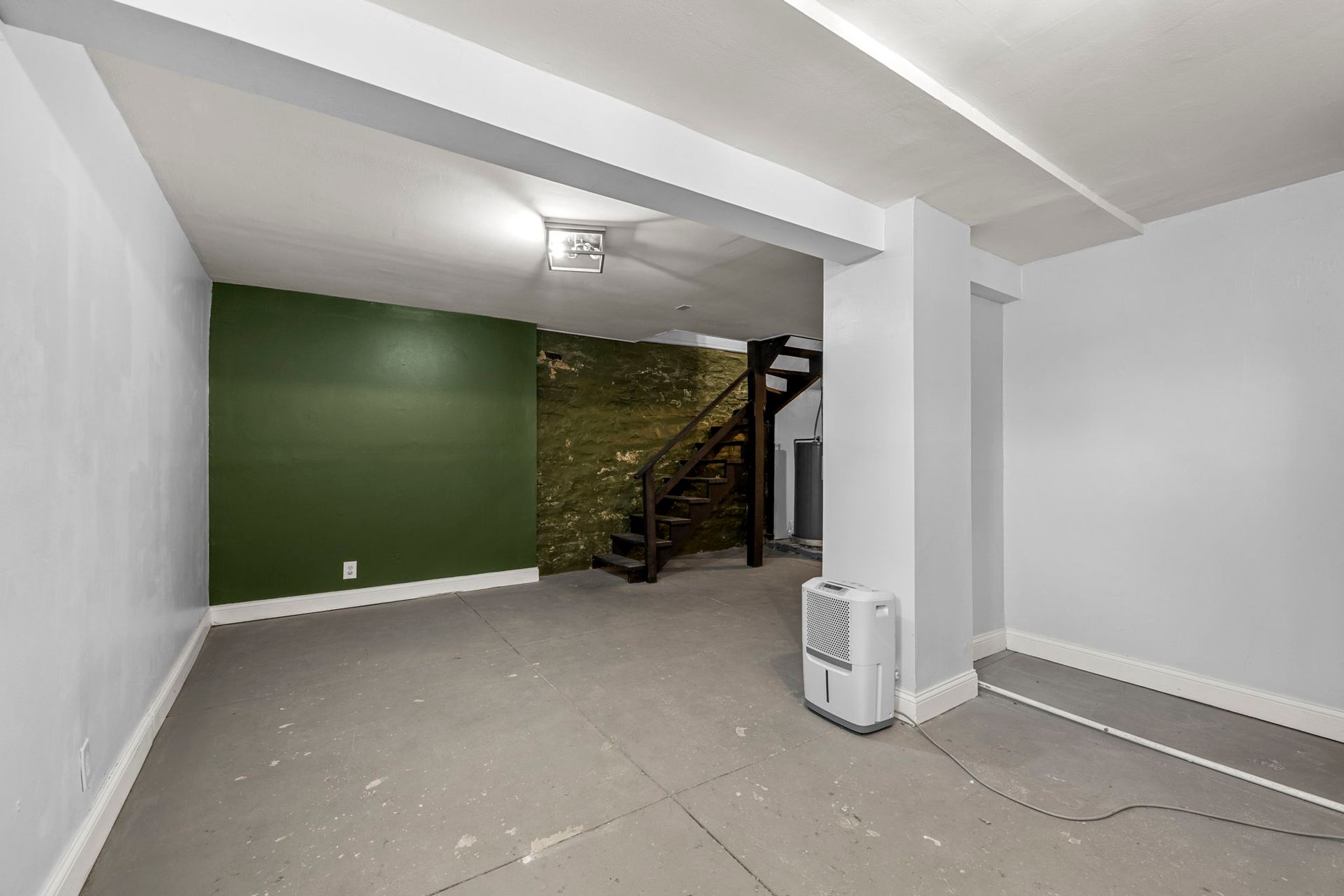 An empty, unfinished room with gray floors, white walls, and a green accent wall. Stairs lead upwards.