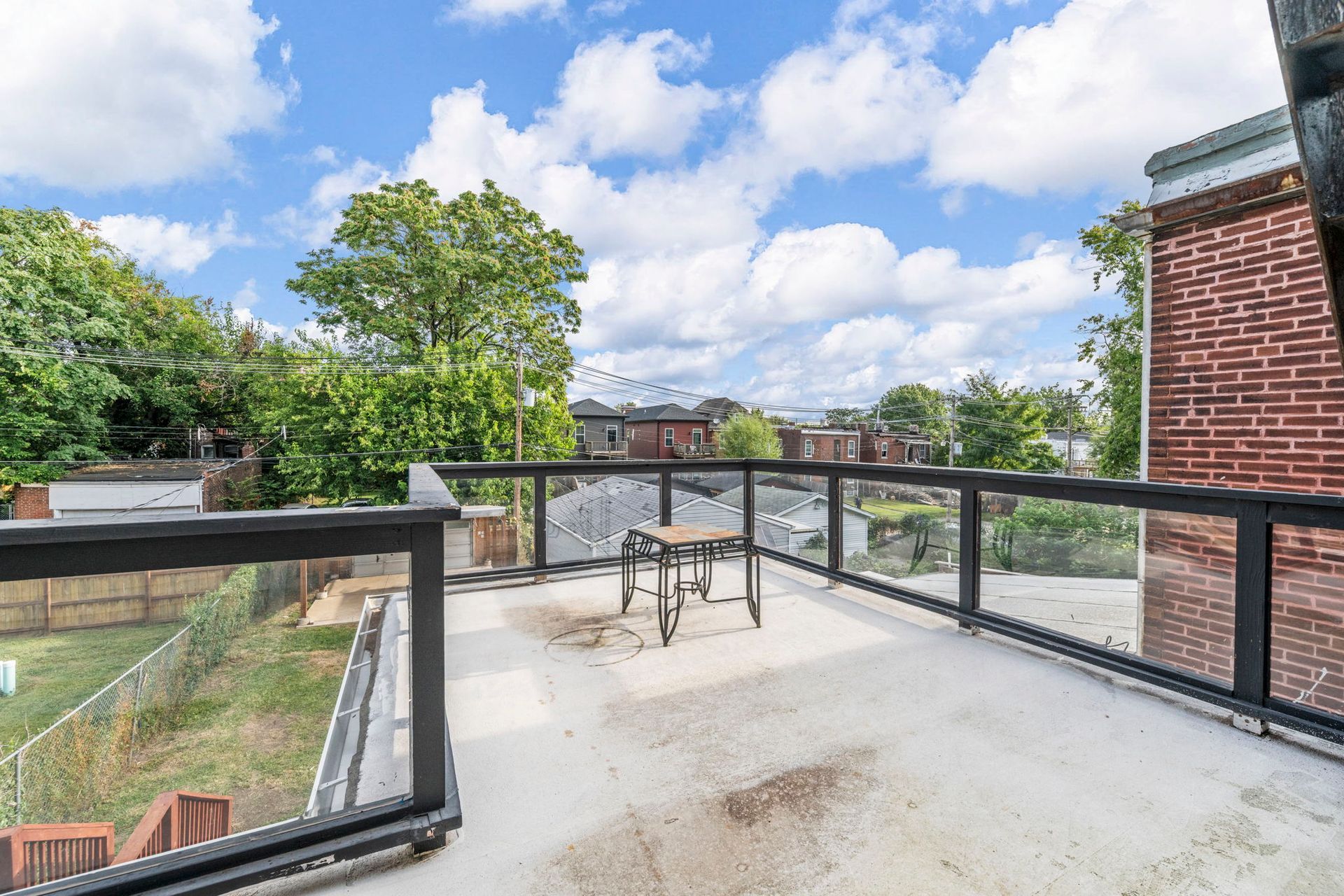 Rooftop deck with glass railing. Overlooks trees, houses, and a cloudy blue sky. A small chair sits on the deck.