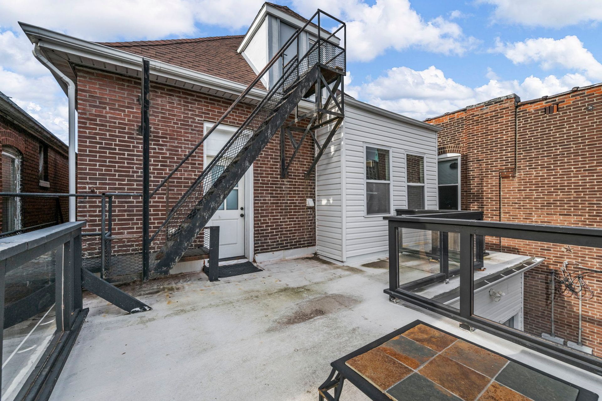 Rooftop with a metal staircase, brick building, white building, and small deck with a table. Cloudy sky.