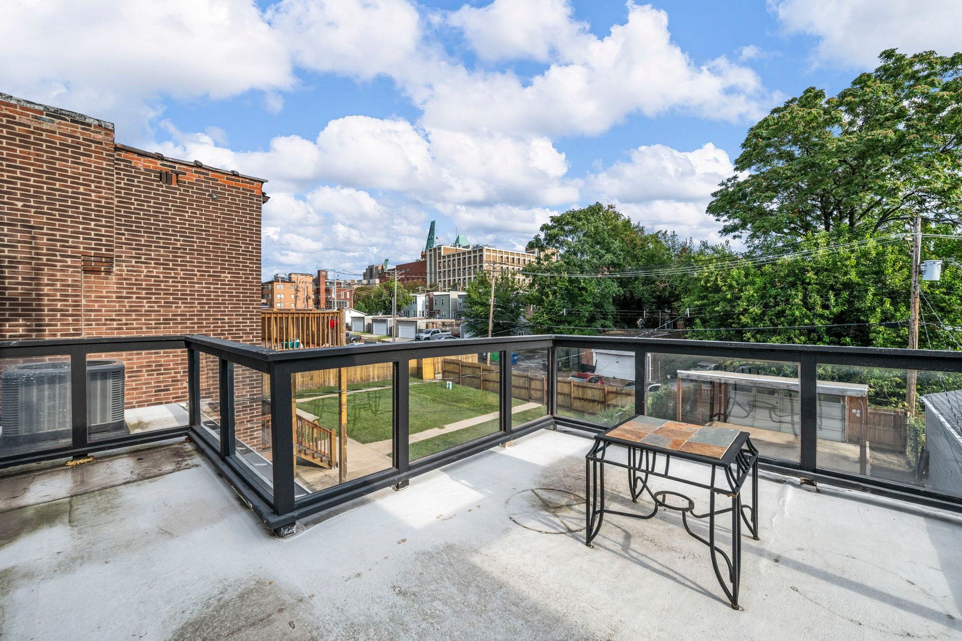 Rooftop patio with glass railings, wrought-iron table, overlooking a yard, brick building, and trees.