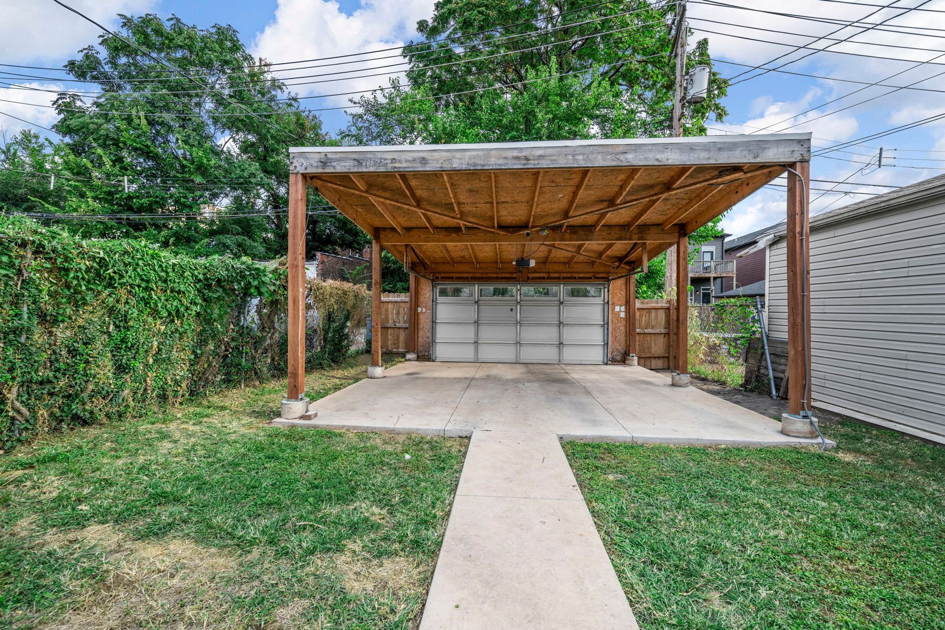 Backyard with a garage and carport. Green grass, a concrete path, and a wooden structure with a white garage door.