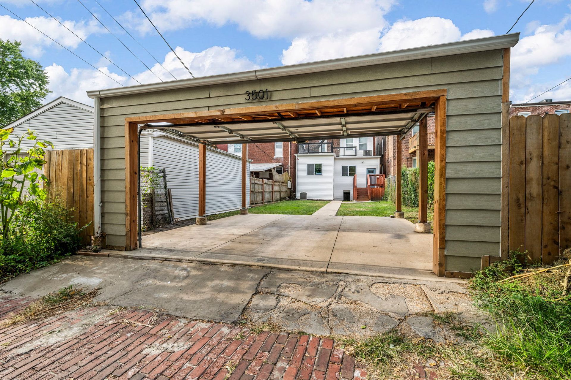 Open-air carport with concrete and brick ground, leading to a backyard with a small house and lawn.