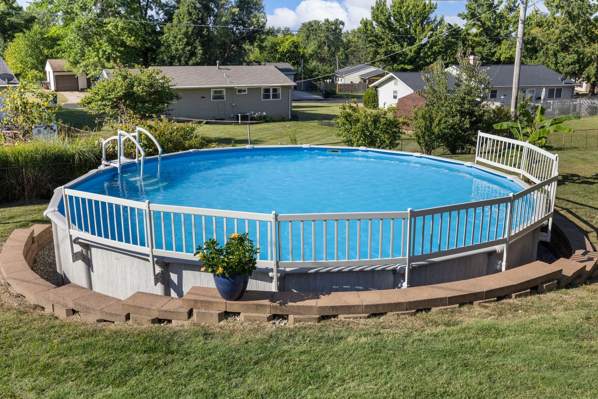 Round above-ground pool with white railing in a grassy yard, surrounded by a brick border, with houses in the background.