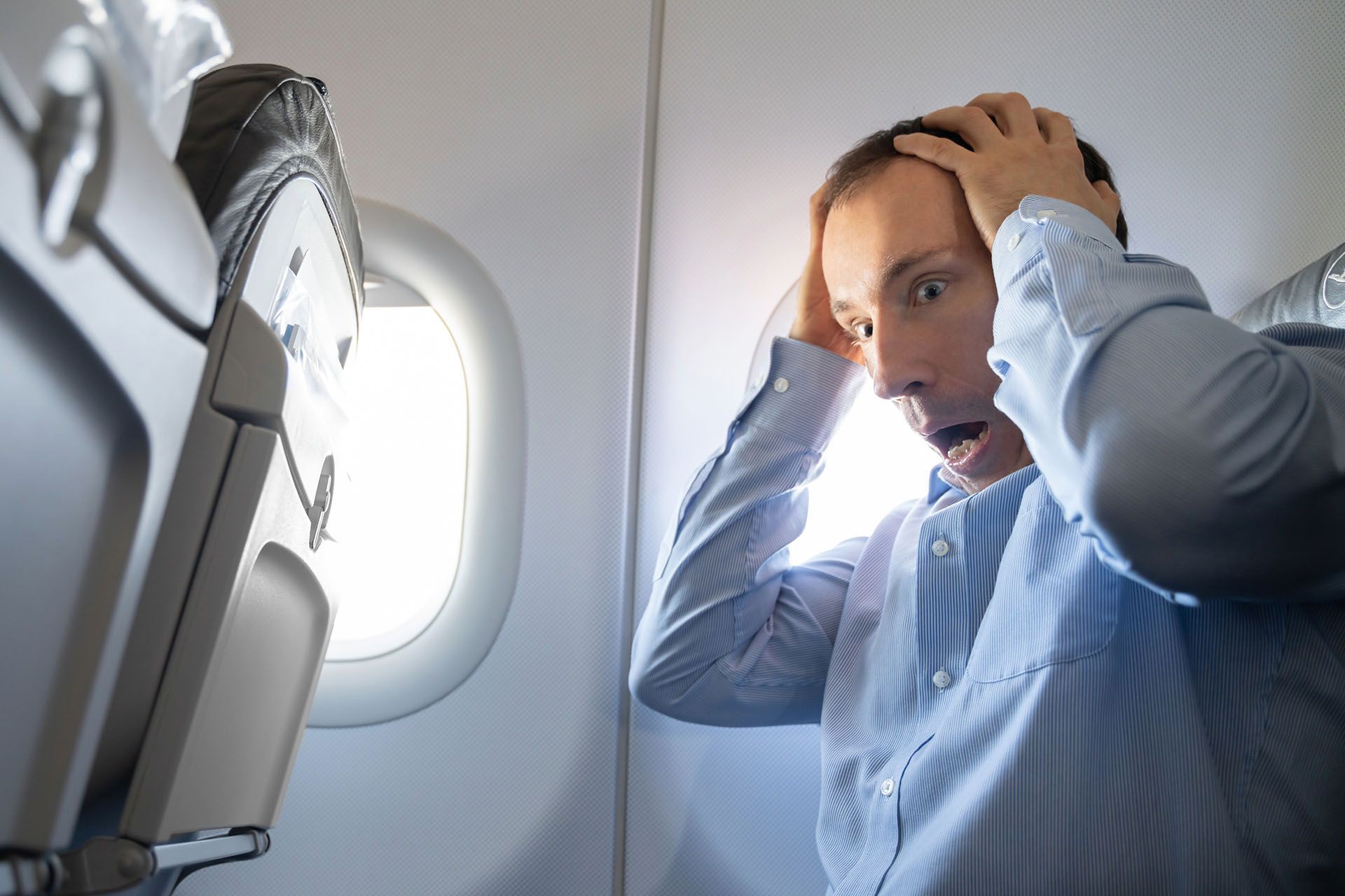 A person sits in an airplane seat, hands on their head, appearing shocked while looking toward a bright cabin window.
