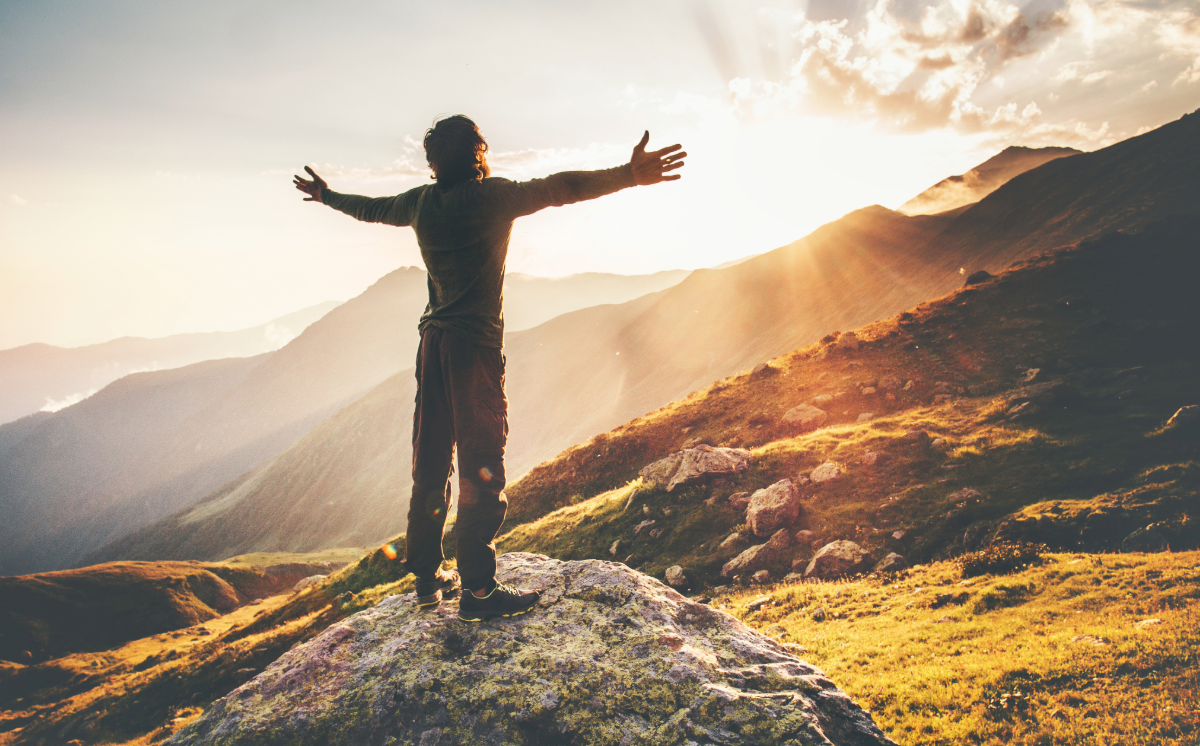 Person standing on a mountain rock at sunrise with arms outstretched over a valley