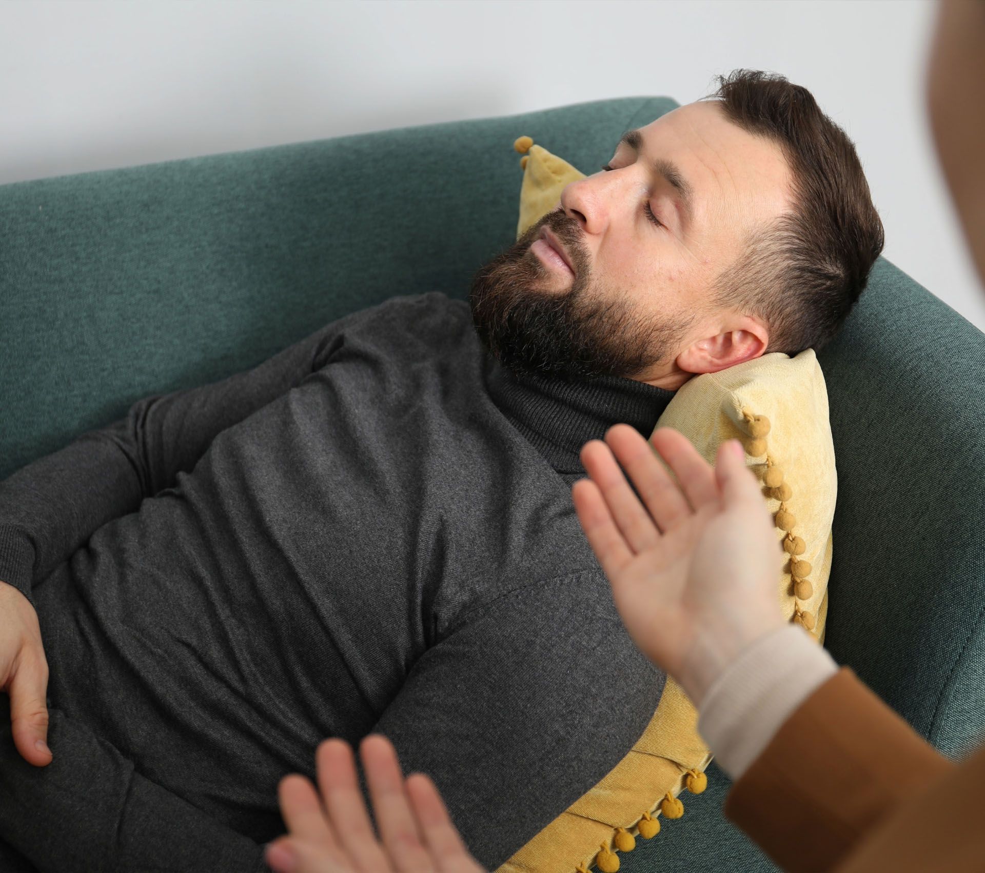 A person lies on a green sofa with eyes closed while someone gestures with their hands during a therapy session.