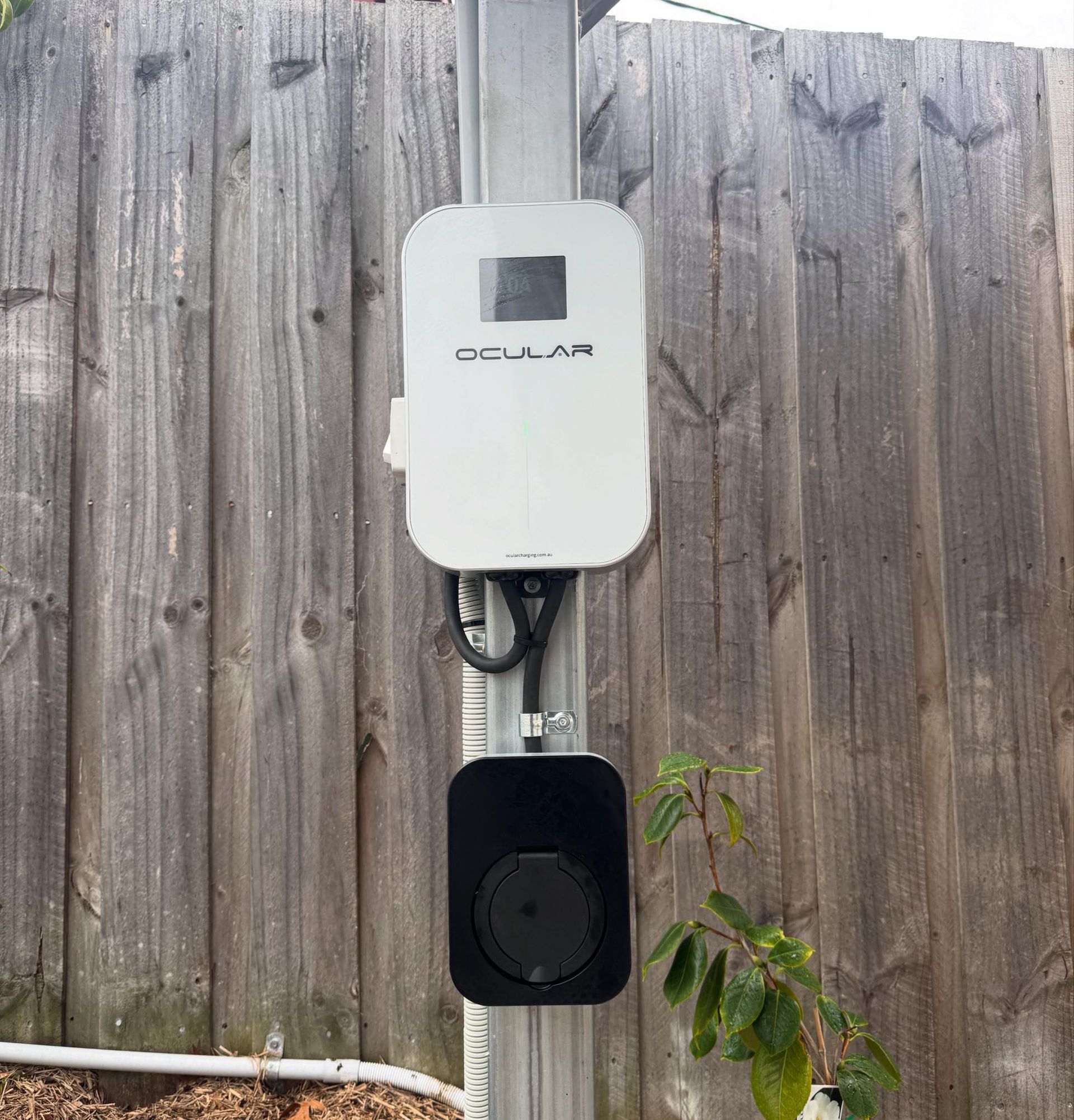 Electric vehicle charger mounted on a post against a wooden fence. The charger is white and black.