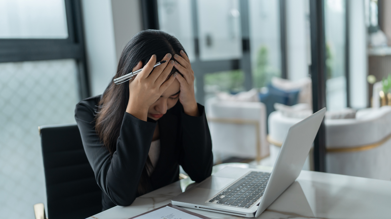 Person at a desk holding their head beside an open laptop in a bright office, looking stressed
