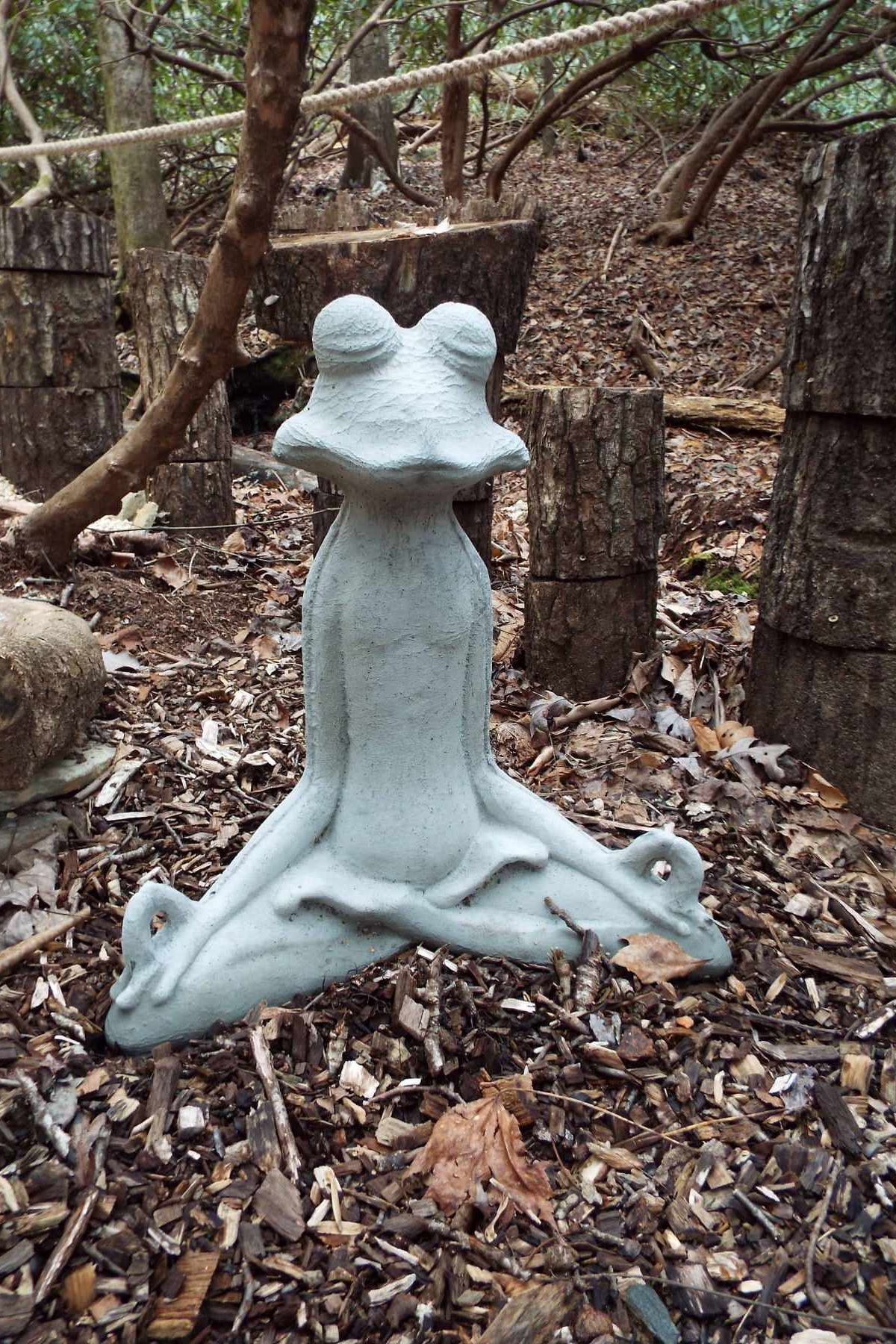 A statue of a frog sitting in a lotus position in the woods.