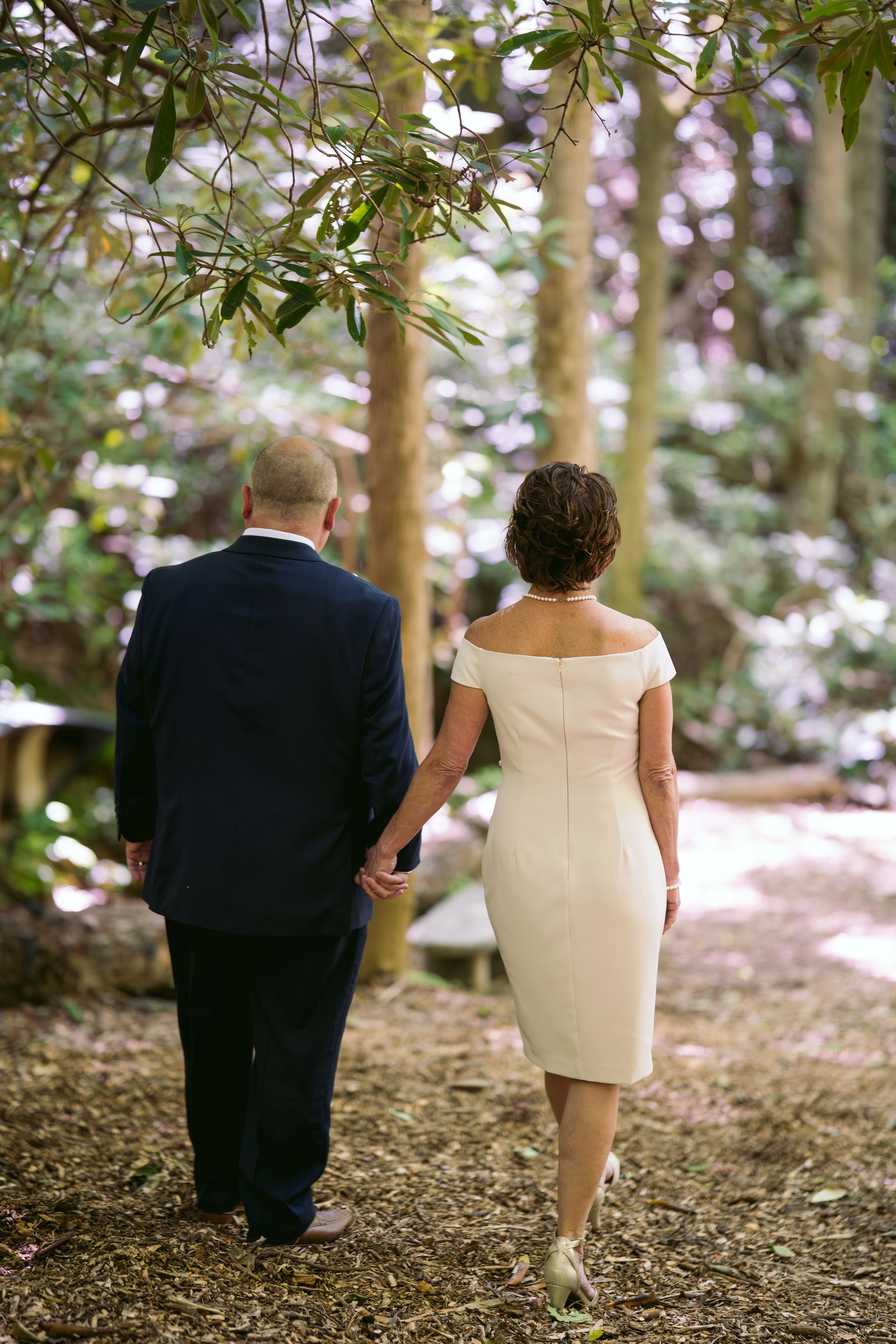 Couple walking hand in hand through a wooded area; the woman in a white dress, the man in a navy suit.