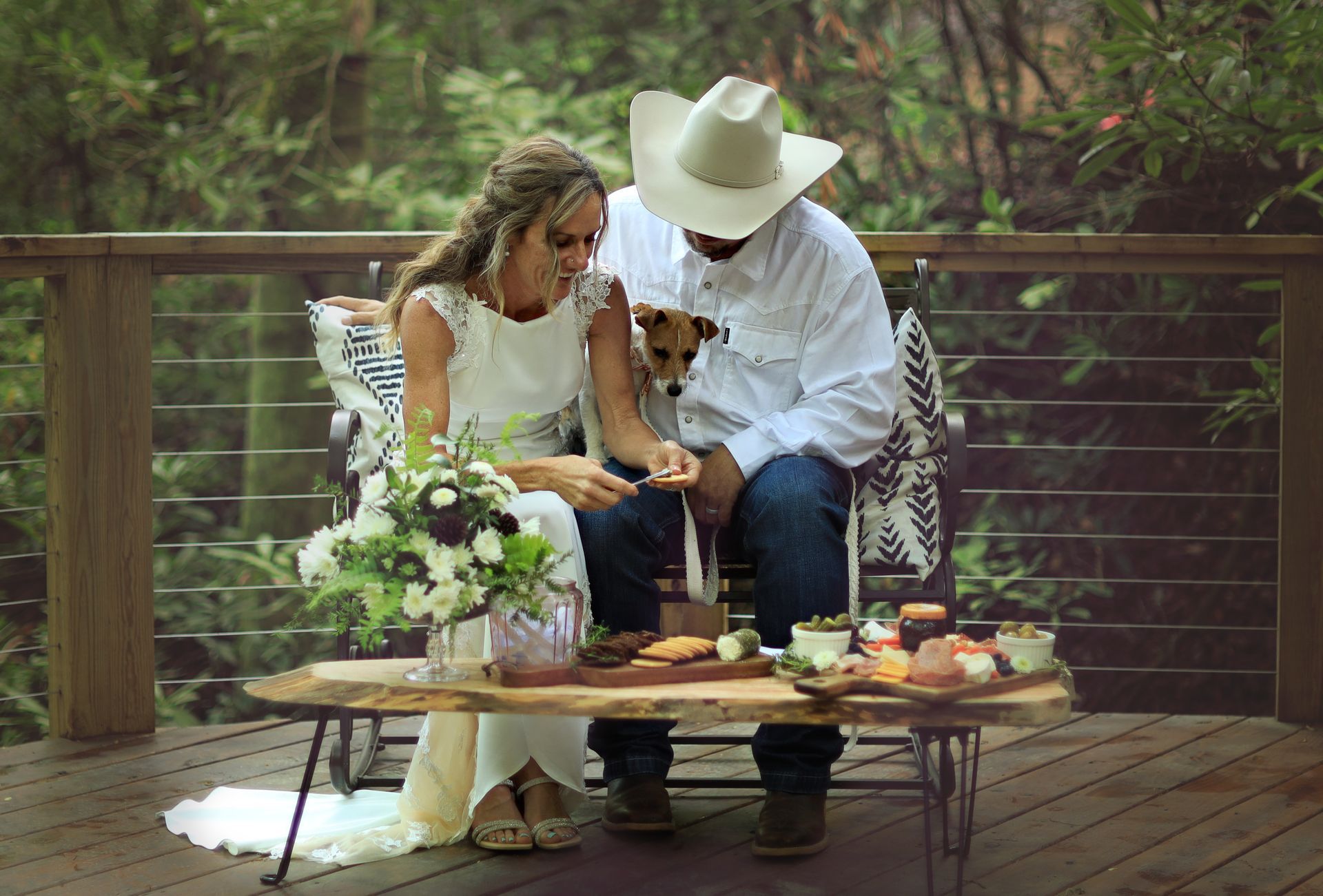 Bride and groom seated on a deck, sharing a wedding cake with flowers nearby