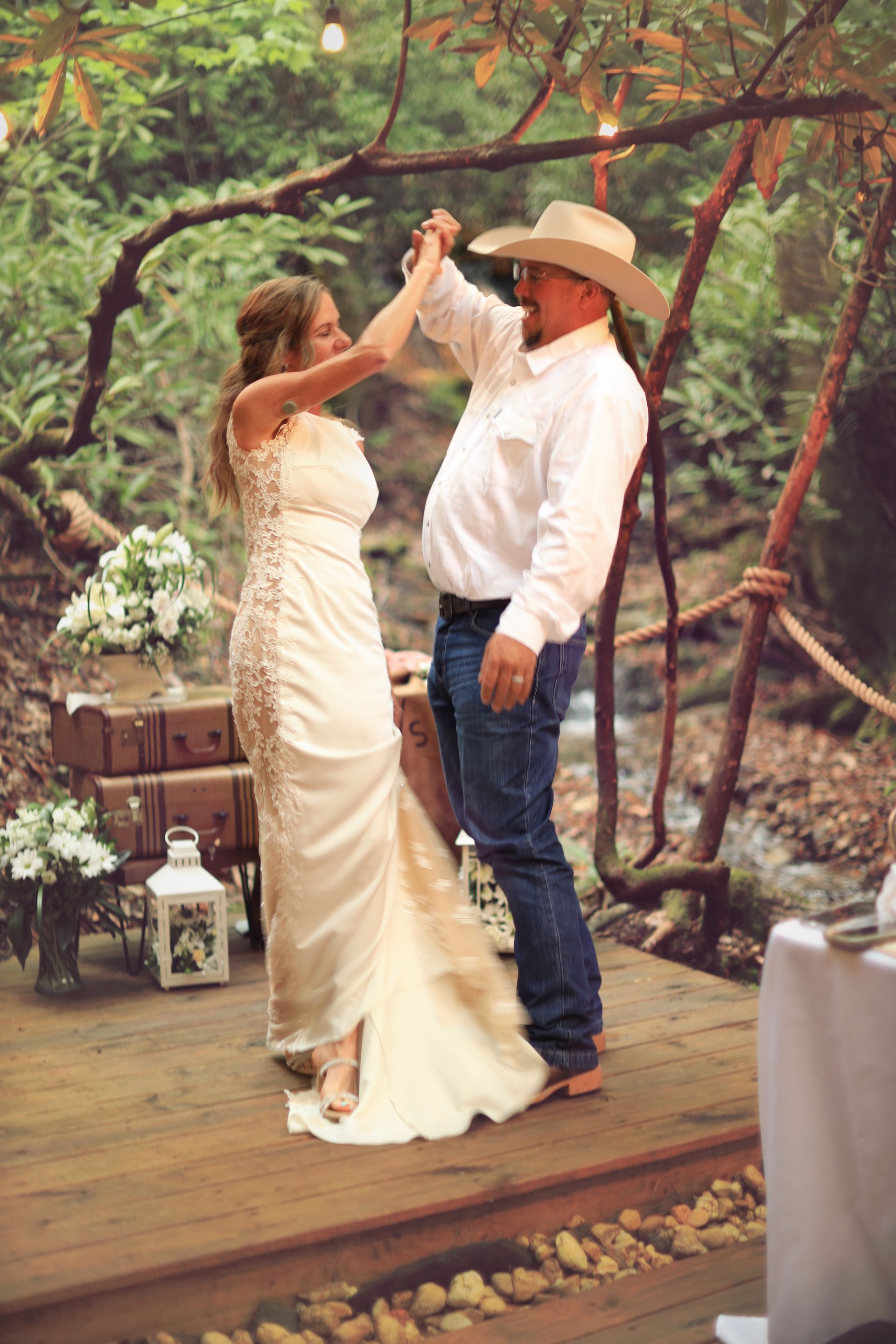 Couple dancing at an outdoor wedding, with a cowboy hat and cream dress under trees.