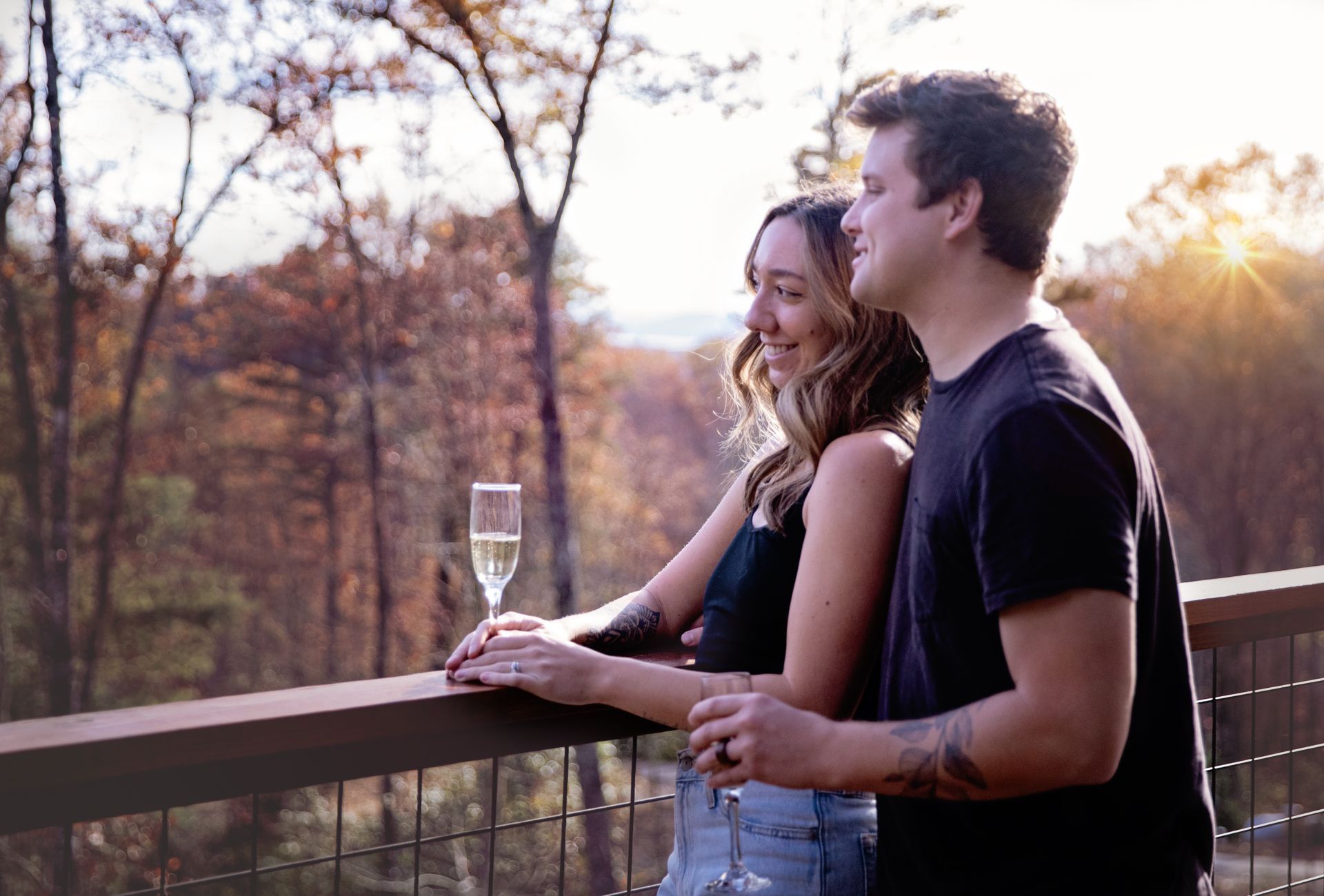 Two people standing on a deck at sunset, holding drinks and looking over a wooded landscape.