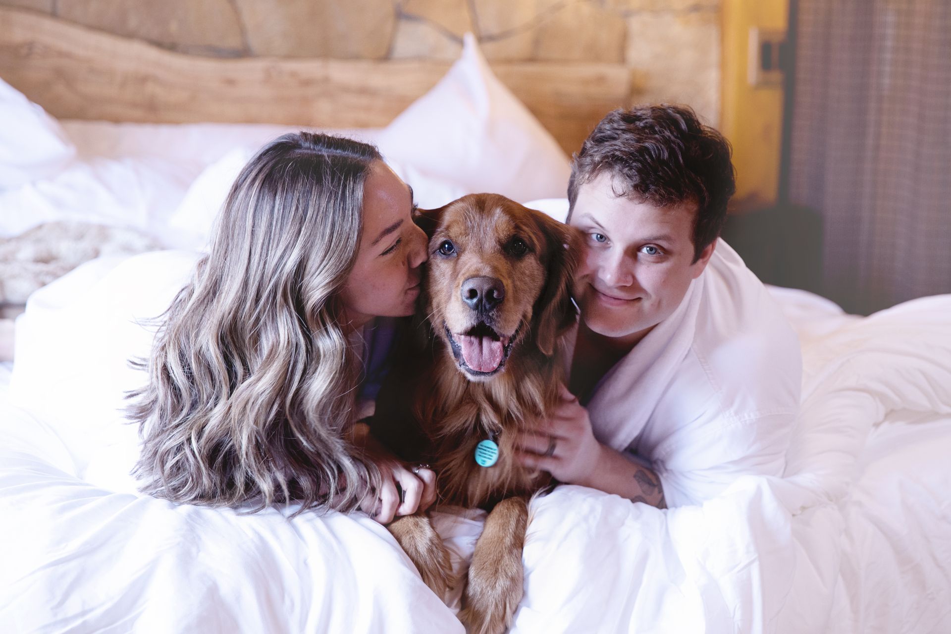 Two people cuddling a brown dog on a white bed, with one person kissing the dog's head