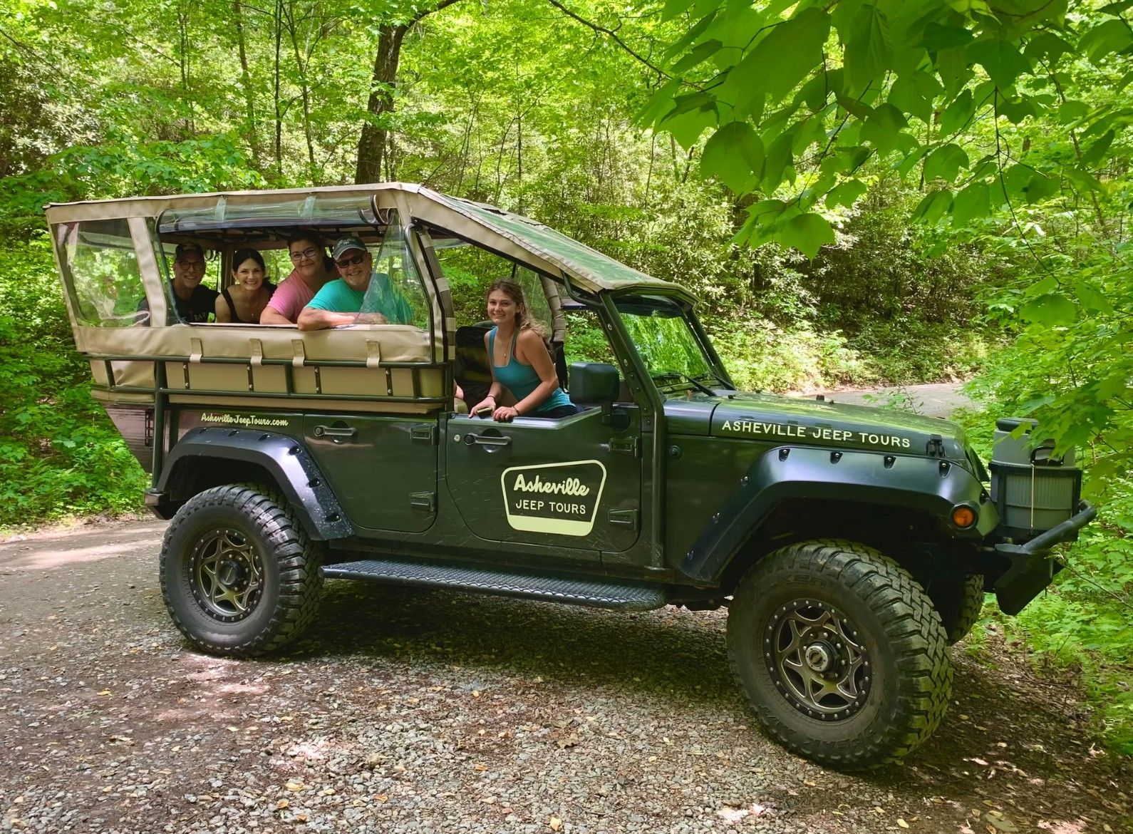 People in a dark green jeep with open sides driving through a lush, green forest.