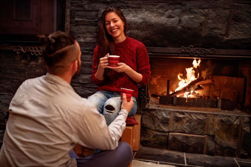 A man and a woman are sitting in front of a fireplace drinking coffee.