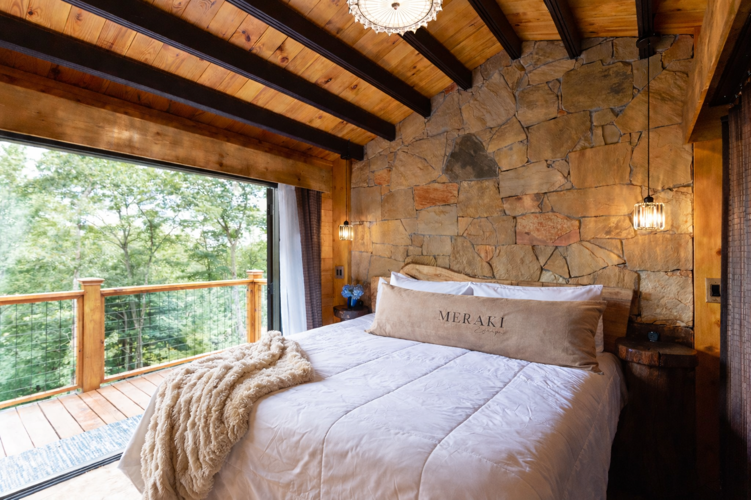 A bedroom with a bed, stone wall, exposed wooden ceiling beams, and a large window looking out onto a wooded balcony.