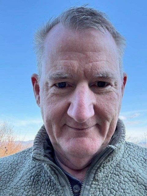 Man with short gray hair and a fleece jacket smiling. Blue sky background.