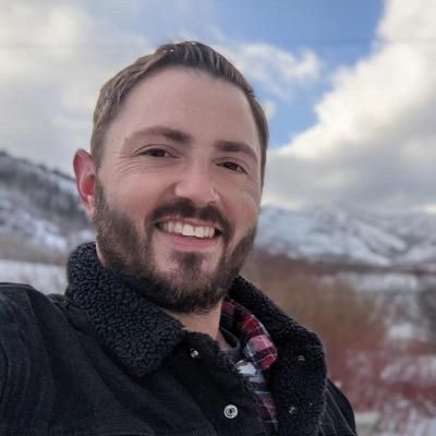 Man smiling, outdoors; wearing black jacket and red plaid shirt, with mountains in the background.
