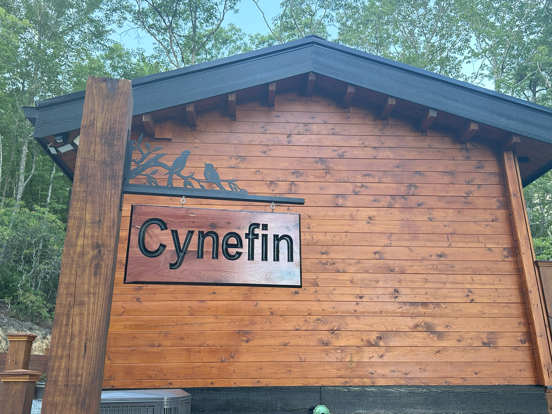 Wooden building with a sign reading “Cynefin” under a dark roof, surrounded by trees.