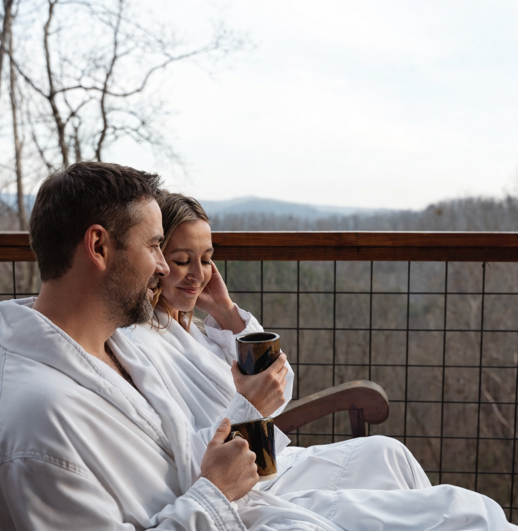 Two people wearing white robes sit on a balcony holding mugs, overlooking a forest landscape during the day.