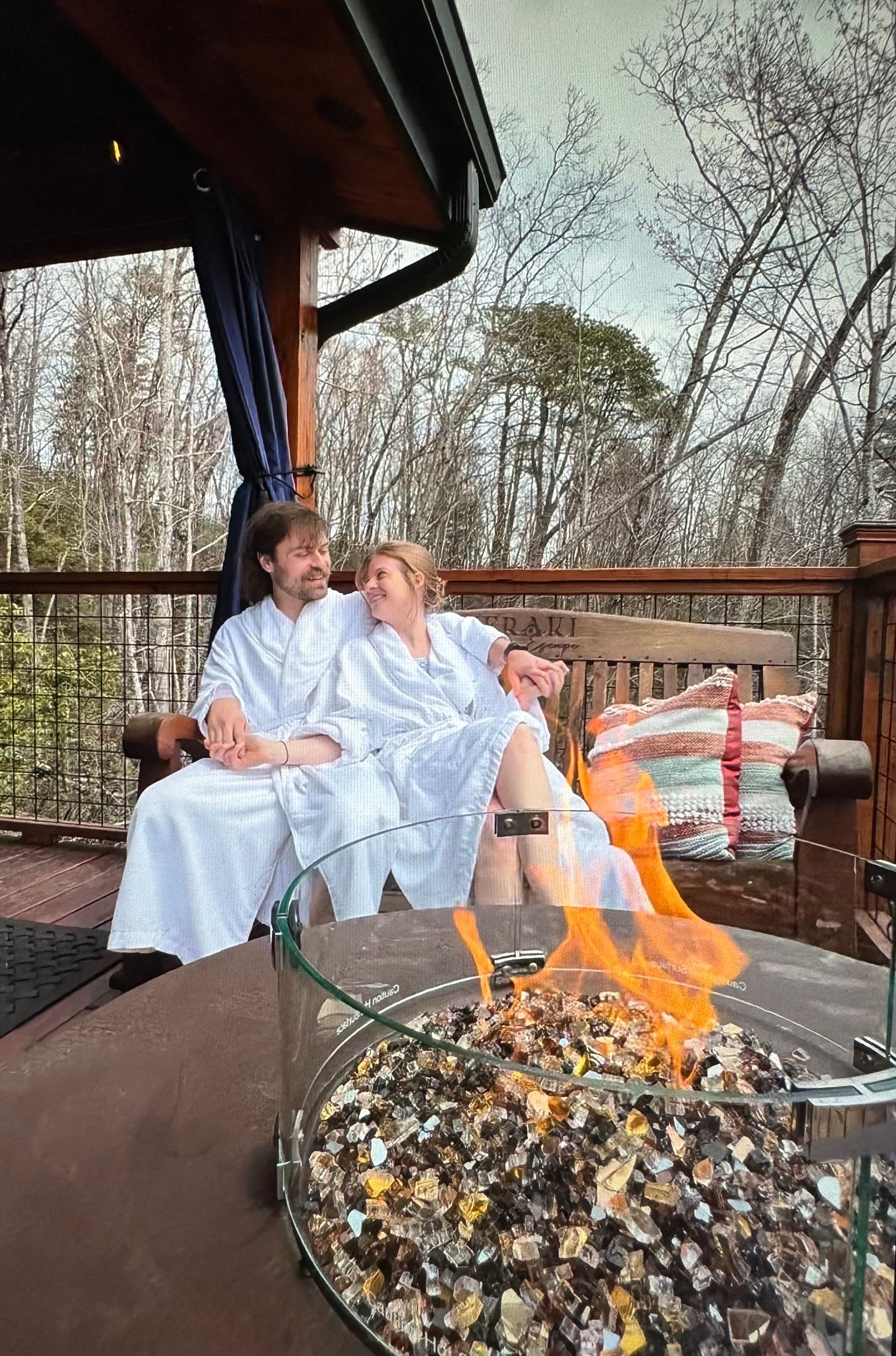 Couple in white robes on deck, looking at each other near fire pit, trees in background.