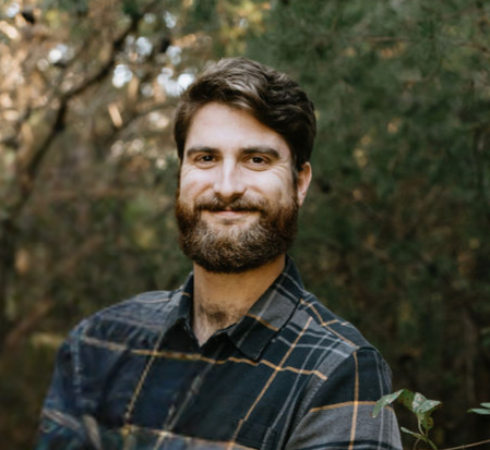 Man with beard smiling, wearing plaid shirt outdoors against a blurry green background.