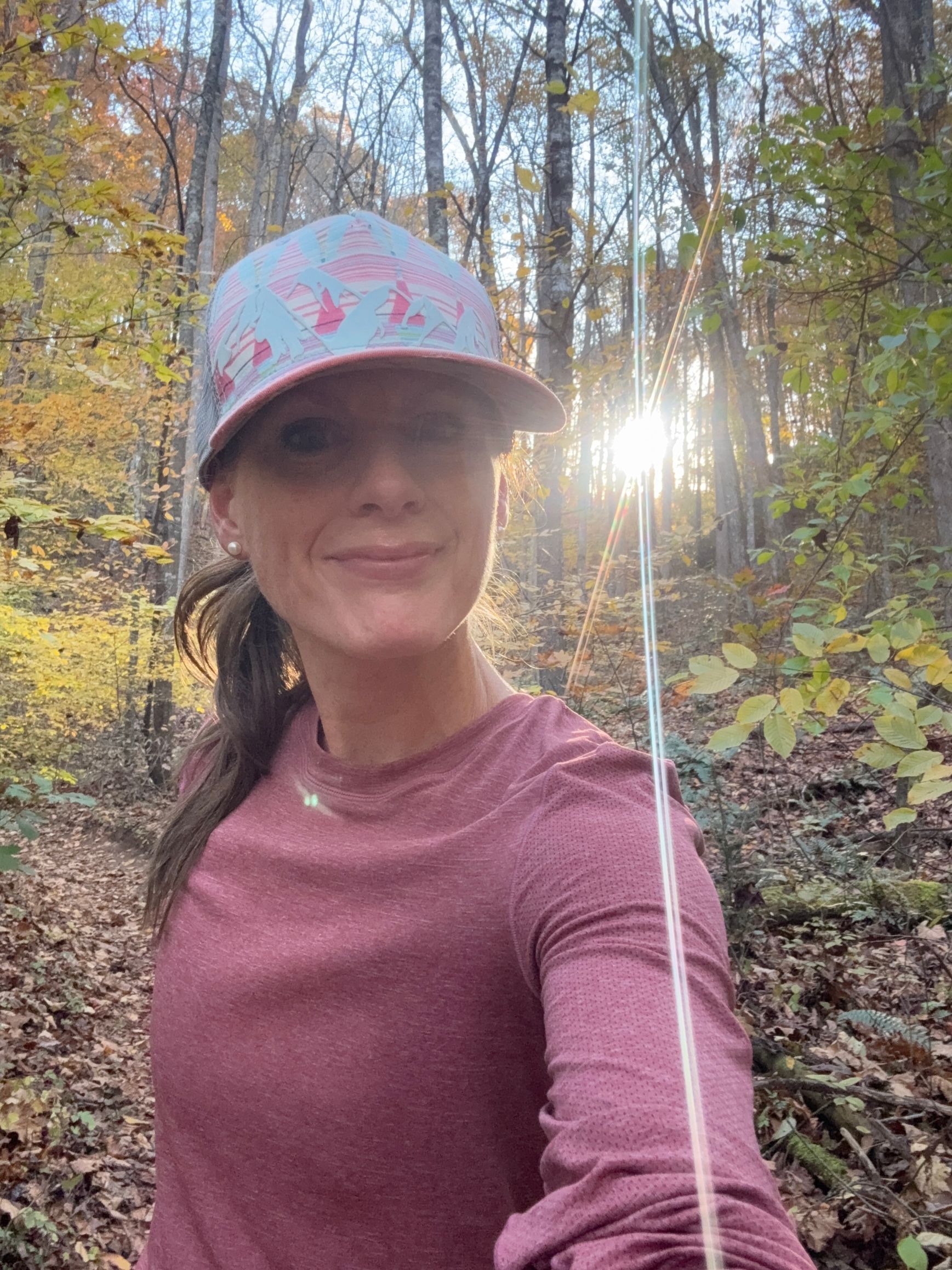 Woman wearing a hat and long-sleeved shirt smiles in a sunlit forest.