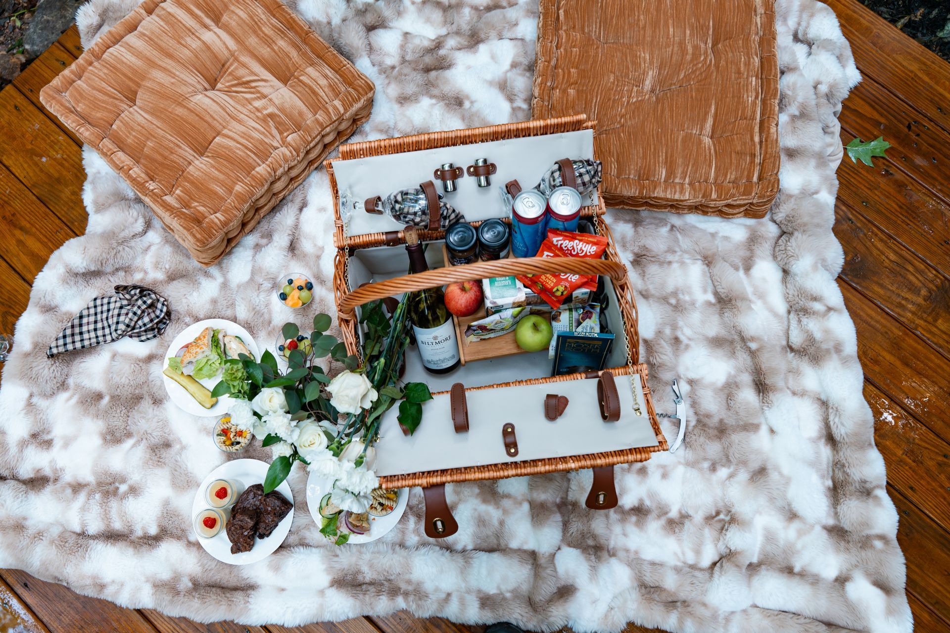 Picnic spread on a blanket beside a forest waterfall, with fruit, pastries, and a wicker basket