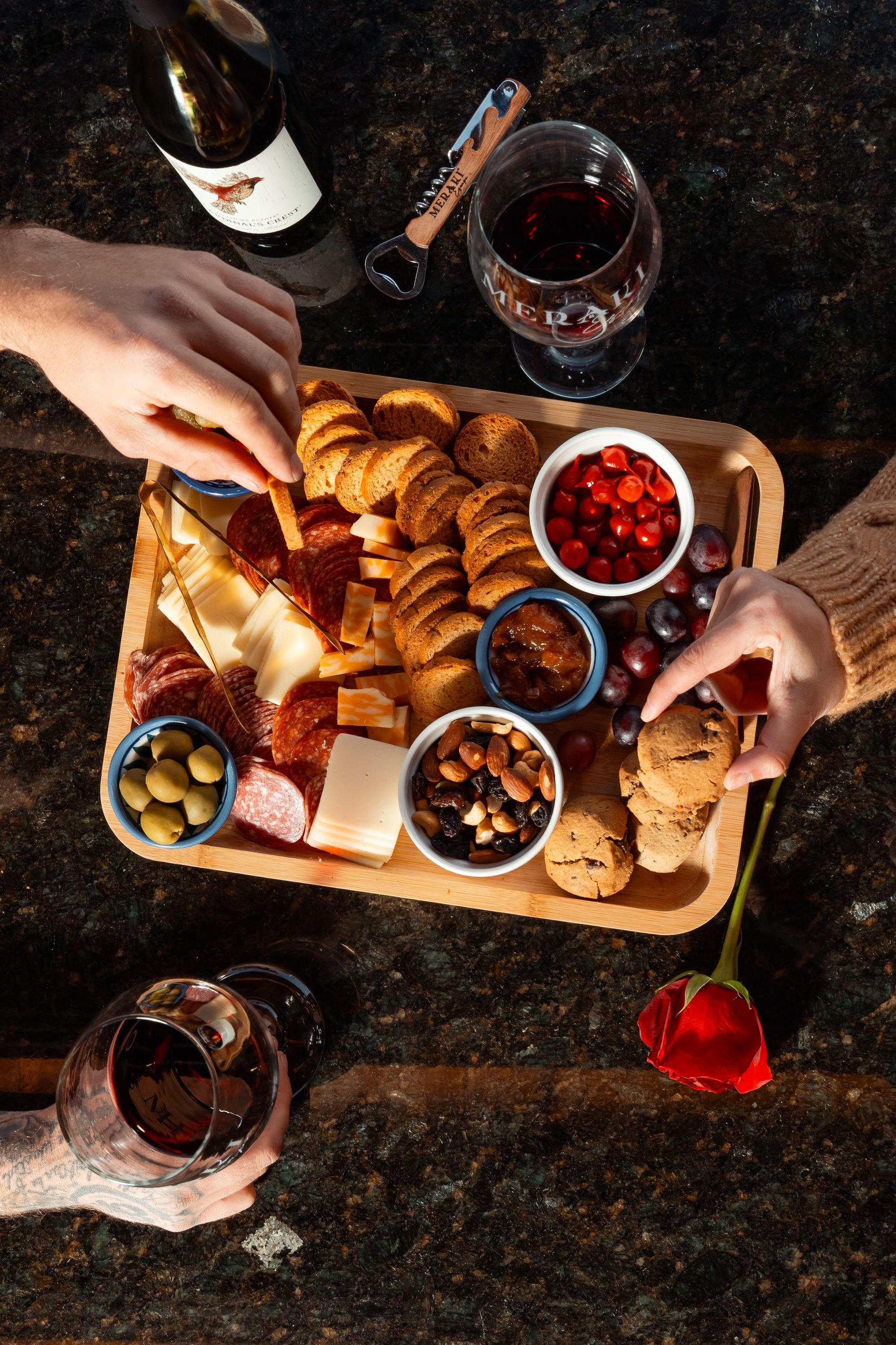 Top-down view of croissants and fruit on a wooden board with wine glasses and hands reaching in