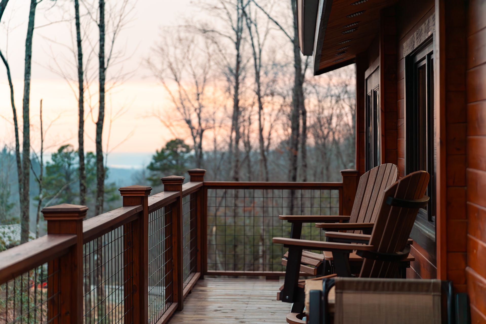 Wooden porch with Adirondack chairs overlooking a bare wooded landscape at sunset.