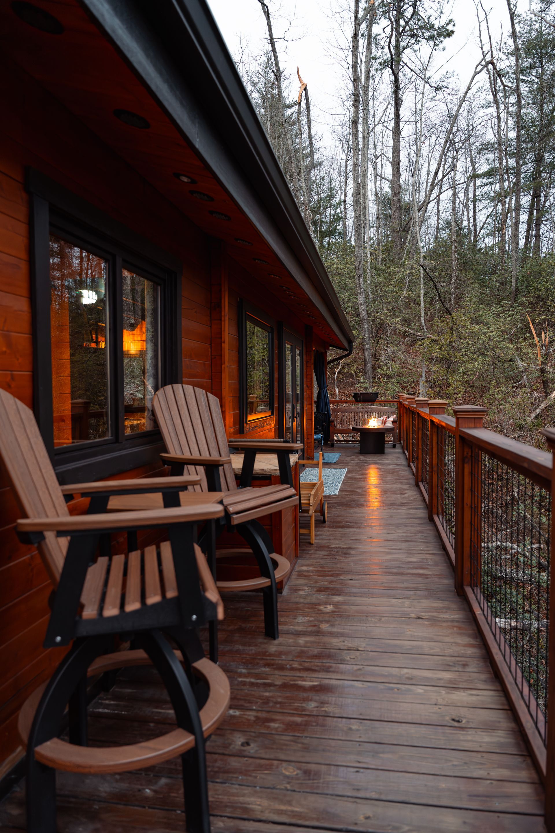 Wooden cabin deck at dusk with chairs, railing, and warm lights overlooking trees