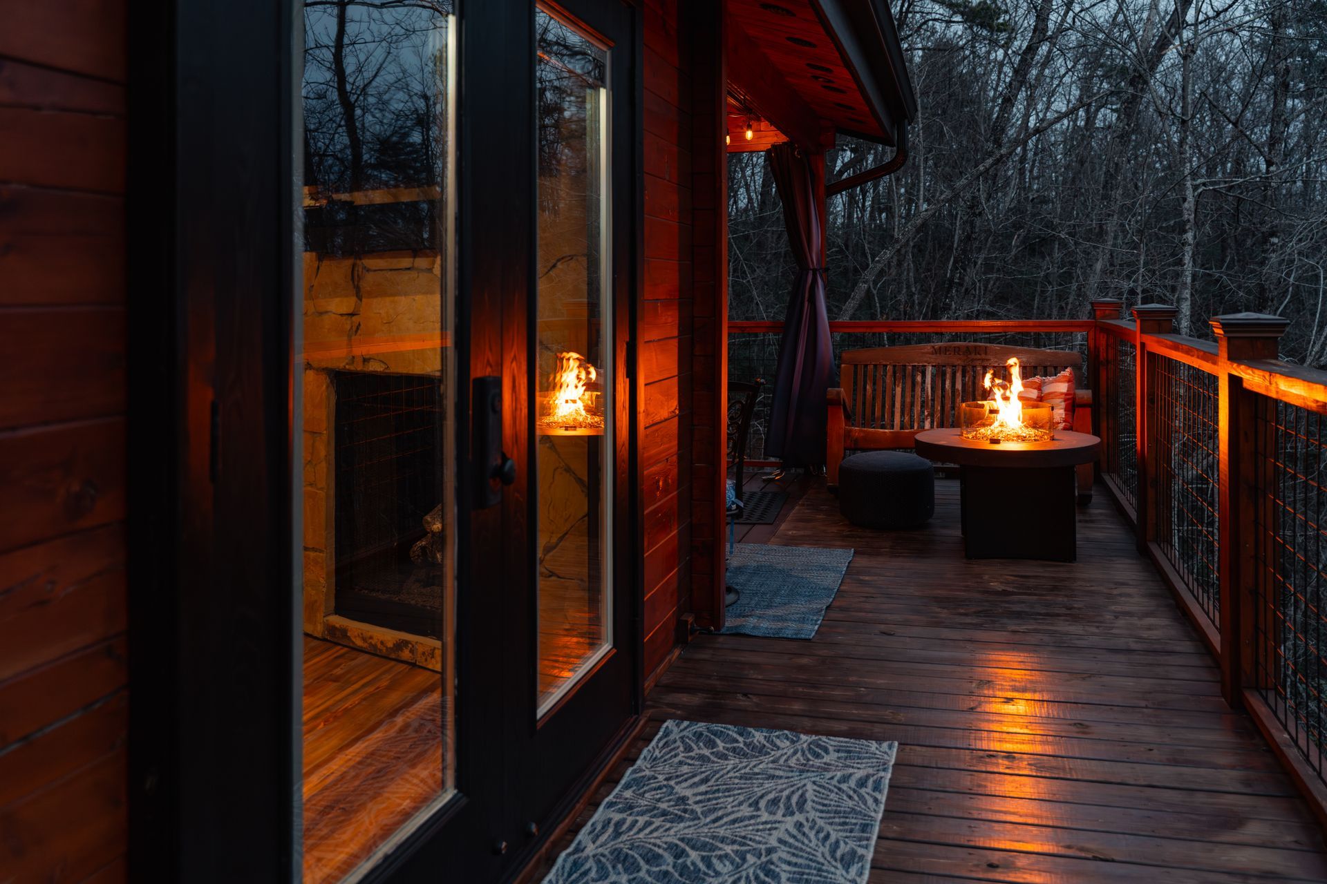 Wooden deck at dusk with warm lanterns, glass doors, and a fire pit by the railing.