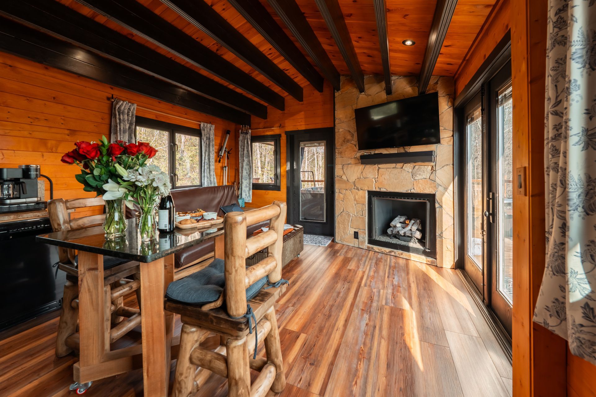 Rustic wood cabin dining area with fireplace, large windows, and a glass table with flowers