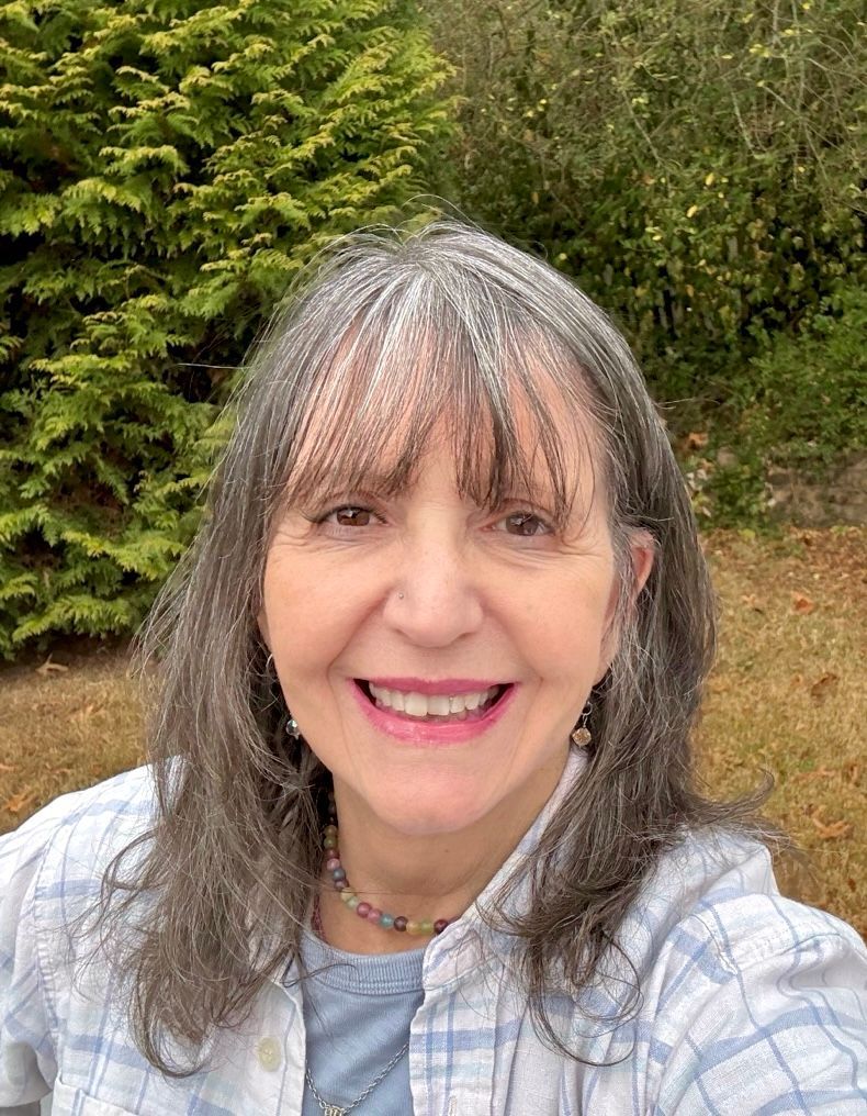 Woman with gray hair and bangs smiles in front of green foliage, wearing a plaid shirt.