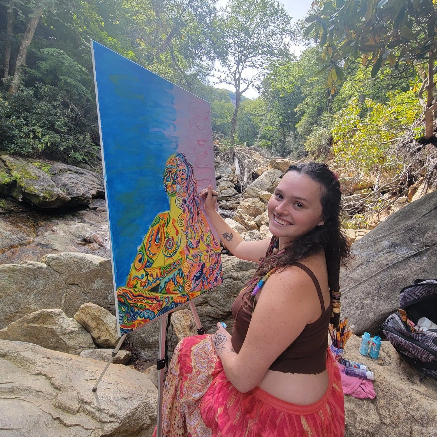 Woman painting on a canvas outdoors, sitting by a stream with a colorful skirt, smiles at the camera.