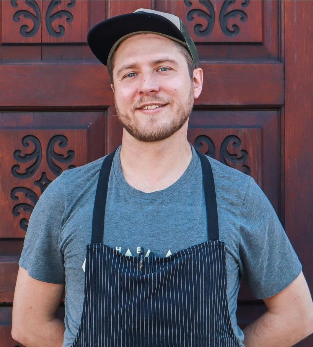 Man in a black cap and striped apron smiling in front of a red wooden door