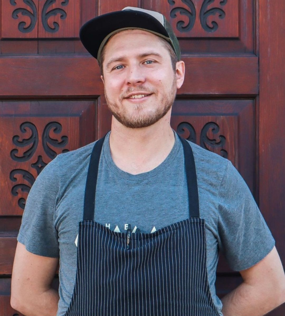 Smiling person in a black cap and striped apron standing in front of a red wooden door