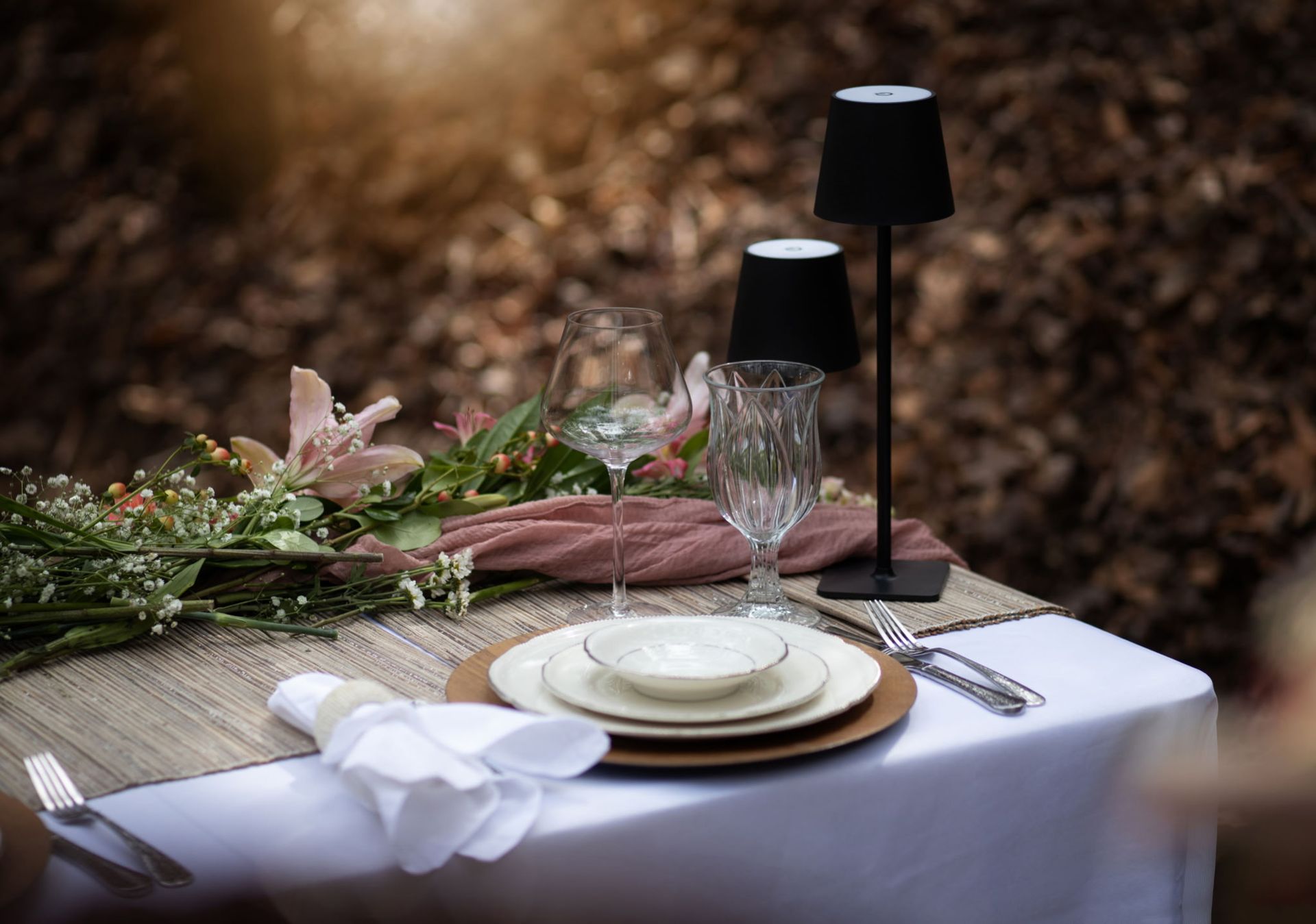 Table setting outdoors with flowers, wine glasses, plates, and black lamps.