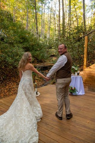 Bride and groom kissing outdoors at wedding ceremony, on a wooden bridge, surrounded by foliage and decorations.