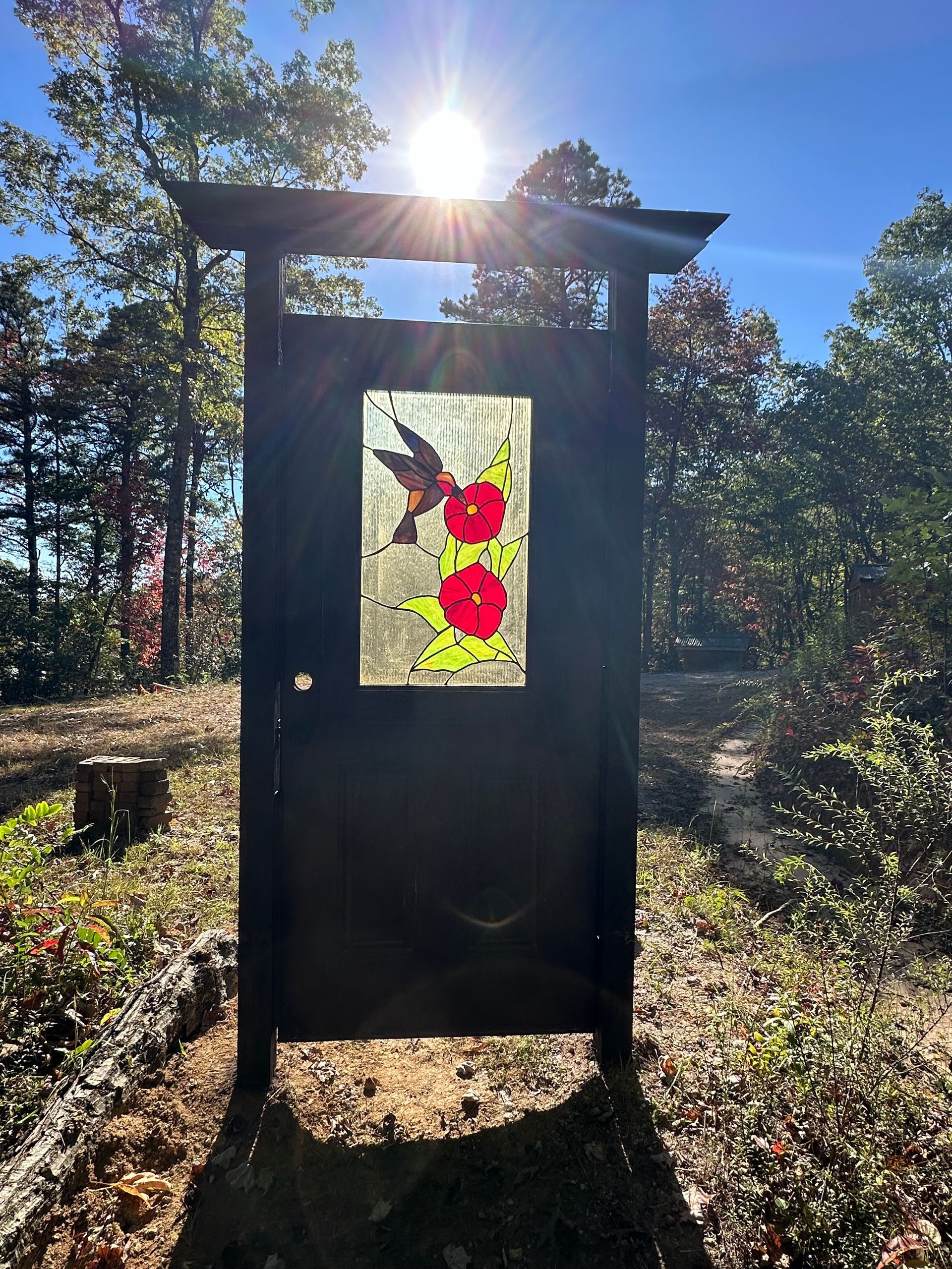 Black outdoor display board in a sunny wooded clearing, with a colorful abstract panel in the center.