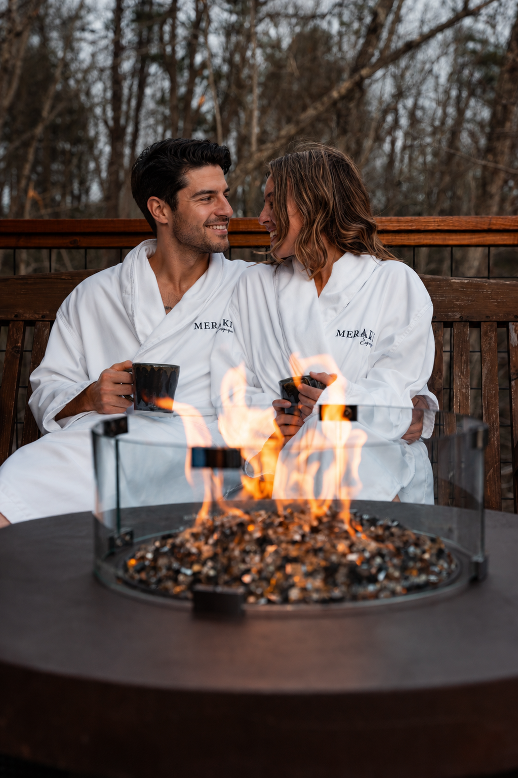 Couple in white robes toasting beside a glowing fire pit on a patio at dusk