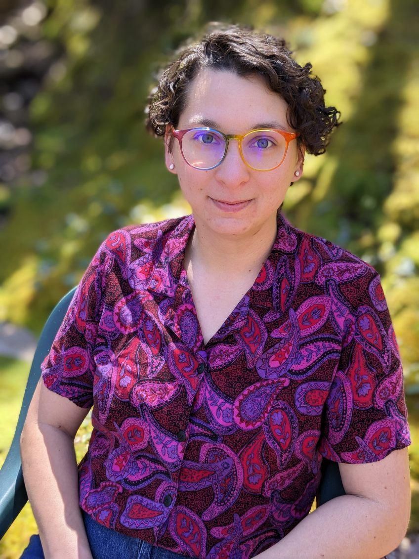 Woman with curly hair and glasses in a purple paisley shirt smiles outdoors.
