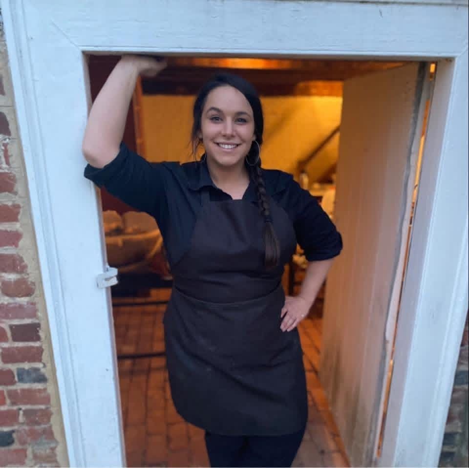 Smiling chef in black apron standing in a doorway with one hand on her hip and one arm raised